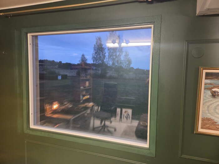 Fönster med grönt foder som matchar väggarna, utsikt över trädgård med träd i skymningen. Tavlor syns i reflektionen i fönstret.
