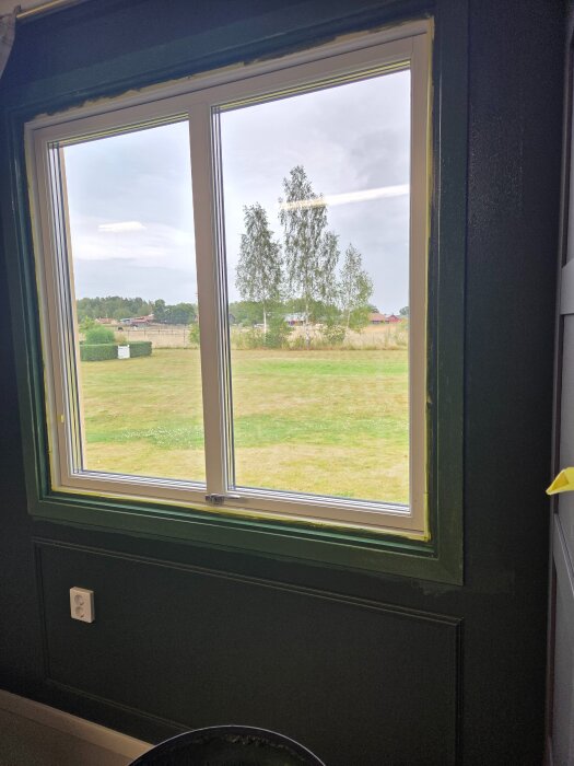 Fönster med nymålade gröna foder som matchar väggarna, utomhusutsikt med gräsmatta och träd syns genom glaset.
