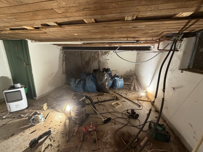 Rum under renovering med synliga rör och balkar i taket, byggmaterial på golvet och upplyst av arbetslampor.