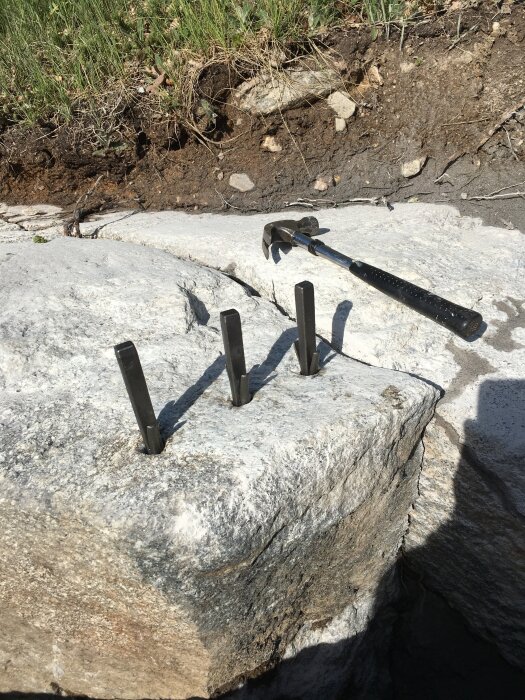 Tre kilar placerade i borrhål på ett stort stenblock med en slägga vid sidan. Bakgrunden består av jord och gräs.