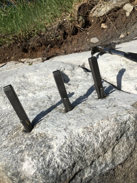 Tre metallkilar sitter i en stor sten som förberedelse för spräckning med hjälp av en hammare, omgiven av jord och gräs.