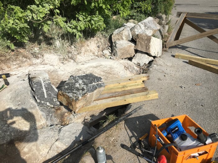 Bilden visar ett spräckt stenblock på en byggarbetsplats med omgivande brädor och verktygslåda, omgiven av naturens grönska i bakgrunden.