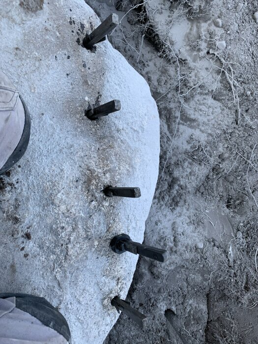 Betongblock med fem utskjutande metallankare på en grusig och torr mark. En del av en skyddssko syns till vänster i bilden.