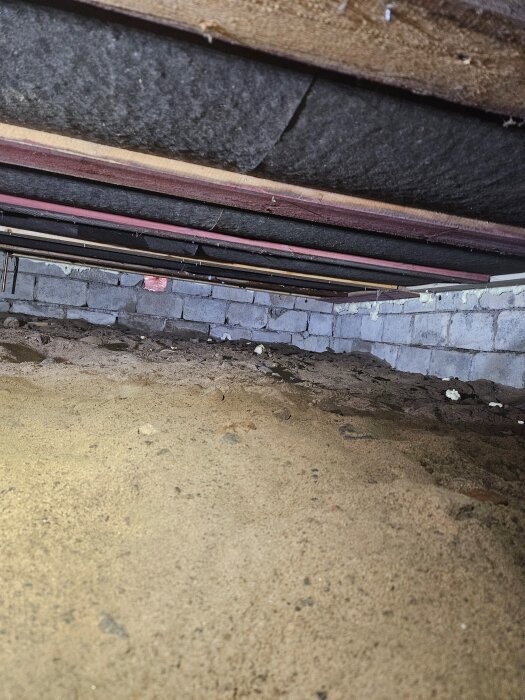 Trång krypgrund med sandgolv och överliggande bjälklag. Blockmur av betongsten syns i bakgrunden med rördragning hängande ovanför.