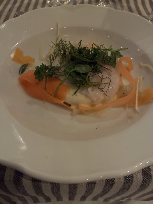 Tallrik med en förrätt bestående av pepparotskräm, färska örter, morot och andra grönsaker, serverad på en randig duk.