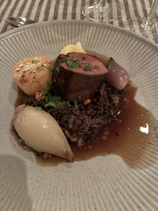En tallrik med köttbit, lök, potatis, linser och sås, garnerad med persilja, serverad på randig duk.