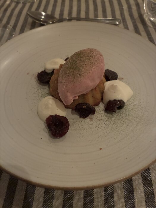Dessert med körsbär, en kula glass och vispgrädde på en vit tallrik, serverad på en randig duk.