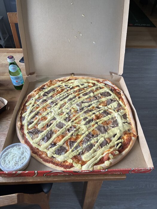 Familjepizza i kartong med vit dressing på toppen bredvid en flaska San Pellegrino och en behållare med vit sallad på bord.