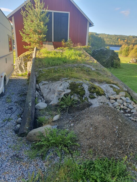 Sten med borrhål i en sluttning framför röd byggnad, omgiven av mossa och grus. Gräs och träd i bakgrunden, vy mot sjö.