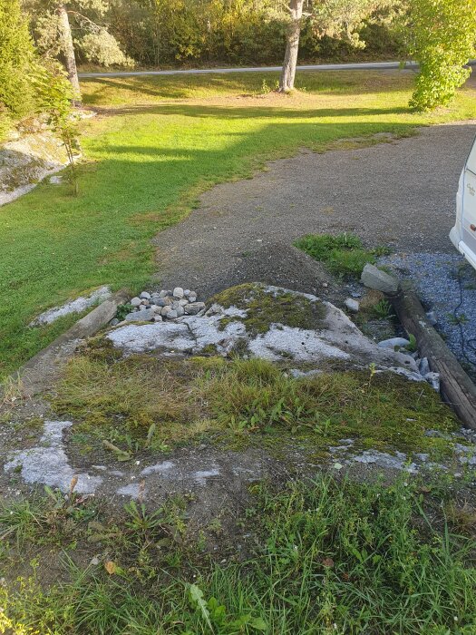 Bilden visar en ojämn stenyta med borrhål och gräs, omgiven av en grusväg och träd, ett område för ett rampbygge.