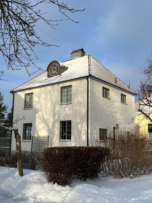 Ett äldre hus, byggt 1918, täckt av snö. Liknar en funkisvilla med vita väggar och brunt tak, omgiven av snötäckta buskar och träd.