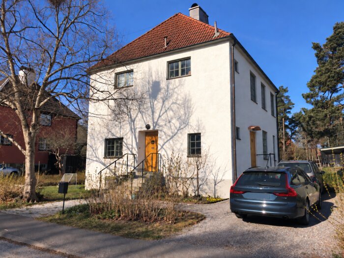 Vit tvåvåningshus med rött tegeltak, solbelyst i en trädgård med parkerad bil, bredvid en röd byggnad och omgivande träd.