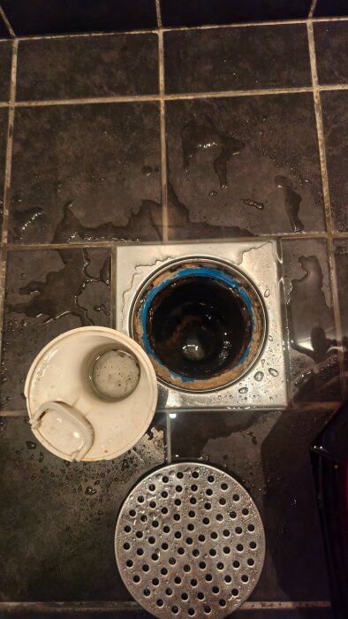 Golvbrunn i dusch med löst vattenlås synligt, inklusiva ett löst lock bredvid. Brunnen har märket Purus. Golvet är blött och kaklat.