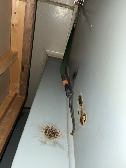 Metallstång med hölje i hörn av ett dolt utrymme, visar brännmärken på väggen. Oklart vad det är, verkar en gång utgjort brandrisk.