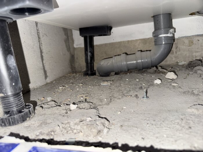Bild av rör under diskho med trasigt betonggolv, exponerade rör och en skruv på ytan.