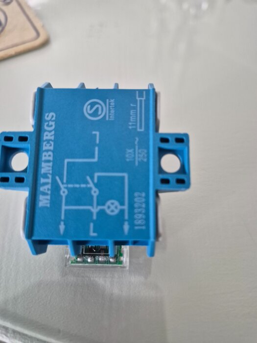 Blå elektrisk komponent märkt "Malmbergs" med tryckt kretsdiagram och tekniska specifikationer synlig ovanpå en yta.