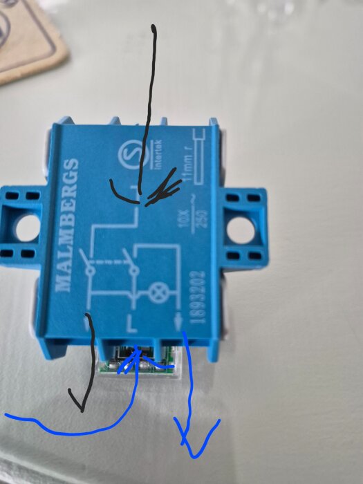 Blå elektrisk kopplingsbox med schematisk diagram och skrift på ovansidan, ritade pilar visar mot olika delar, fotograferad på en ljus yta.