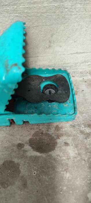 Turkos råttfälla på cementgolv, utan bete, med öppet lock. Används för att fånga möss, trots brist på lockbete.