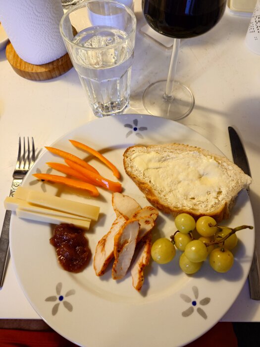 En tallrik med oststavar, kycklingskivor, röd marmelad, orange paprika, vitt bröd och gröna druvor. Ett glas vatten och rödvin i bakgrunden.