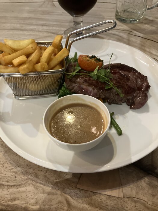 Steak med grillad tomat och grönt, serverad med pommes frites i korg och sås i skål på vit tallrik.