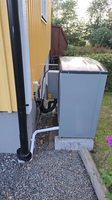 Utomhusmonterad enhet av S2125 luft-vattenvärmepump vid husfasad med gul väggpanel och dräneringsrör på grusunderlag.