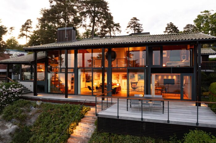 Modern villa med stora glaspartier, upplyst interiör, trädäck och trappa i solnedgång. Omgivande träd och vacker trädgård i förgrunden.