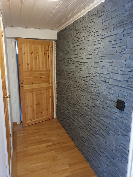 Stenvägg med dold dörr i ett renoverat rum. Vägg av staplade grå stenar och en träfärgad dörr med paneldesign. Trägolv och vitmålat tak.