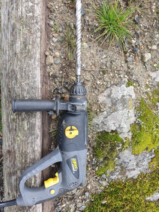 Borrmaskin med borrspets, inställd på slag och hög hastighet. Ligger på mark med grus och mossa.