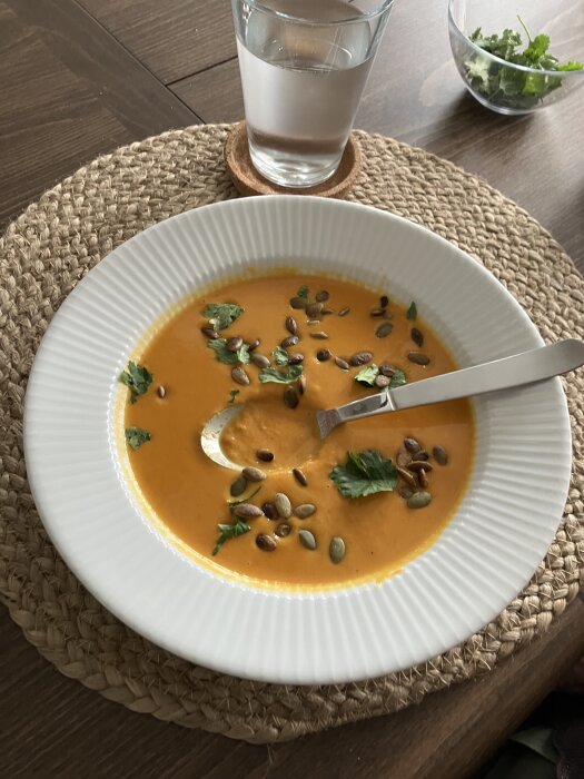 Orange soppa med frön och örter i en vit skål på en flätad underlägg, med ett glas vatten bredvid.