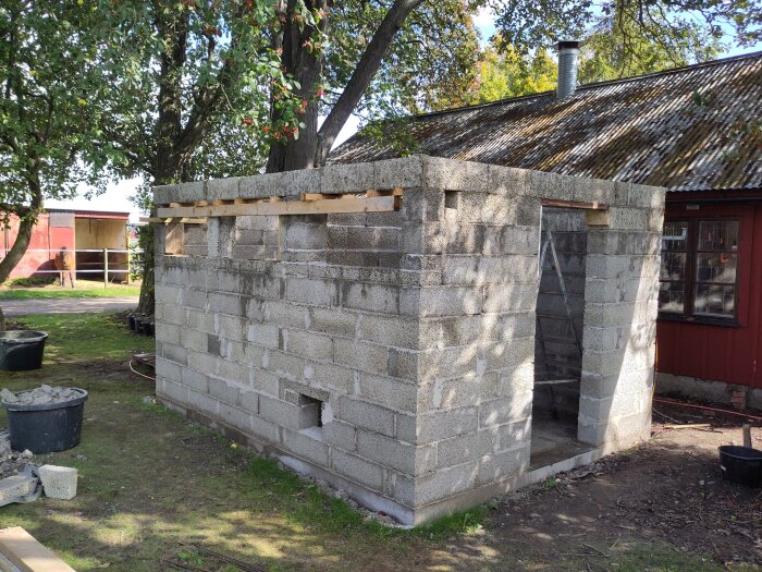 Ouppfört murverk av betongblock intill hus, träreglar på tak, stege inuti. Stora träd i bakgrunden, soligt väder.