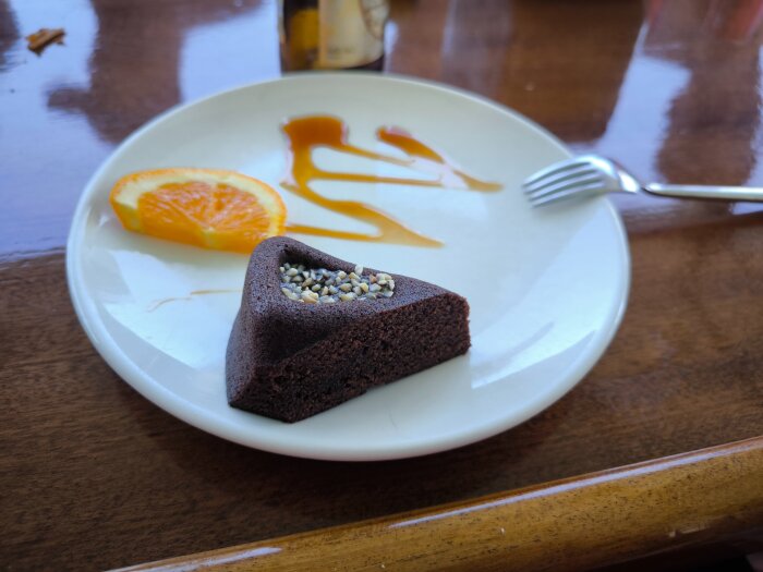 Chokladkaka med nötter, en apelsinskiva och sås på en tallrik, serverad på ett träbord med en gaffel.