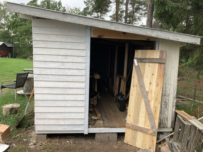 Ett utomhusskjul med öppen dörr, synligt trägolv av trall. Inuti finns ved och ett utrymme för förvaring till höger.