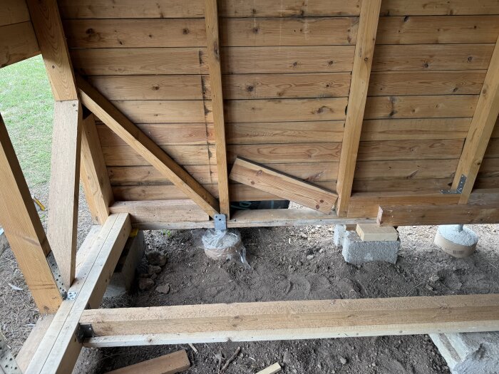 Trävägg på bod byggs upp med nya plintar och reglar. Ny syll monteras under gamla träpaneler, synligt jordgolv och träkonstruktion.