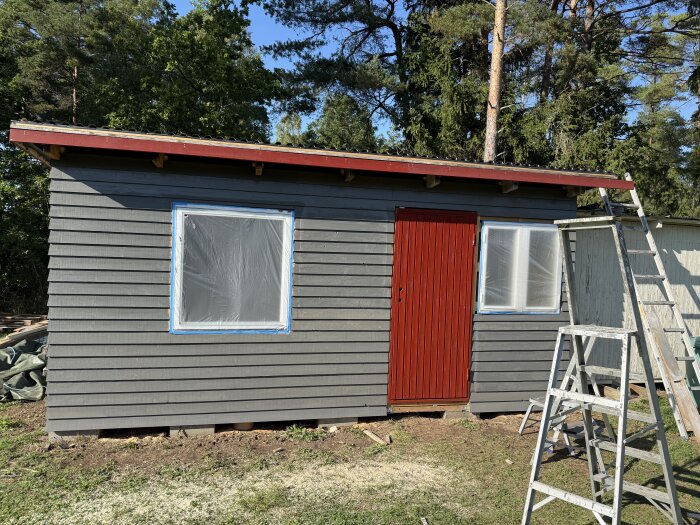 Gråmålad byggnad med fjällpanel, vitt plastskyddat fönster och röd dörr. En stege står bredvid, omgivningen är träd och gräs.