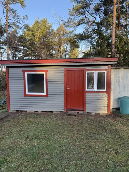 Litet grått skjul med röd dörr och fönsterkarmar, omgiven av träd, beläget på gräs.