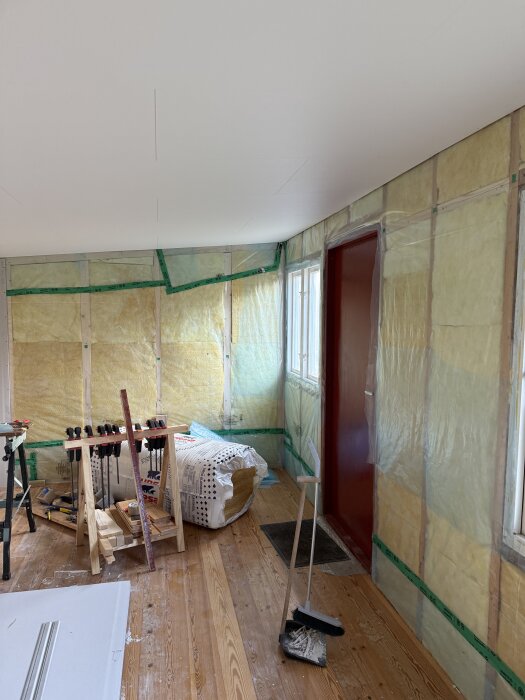 Renovering pågår med träbjälkar, verktyg och isolering längs väggarna. Omonterade saker och byggmaterial på trägolv.