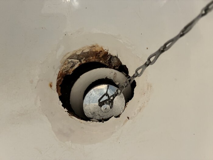 Närbild av en gammal avloppsbrunn med rost och skador i emaljen på ett inbyggt badkar, kedja fäst vid proppen.