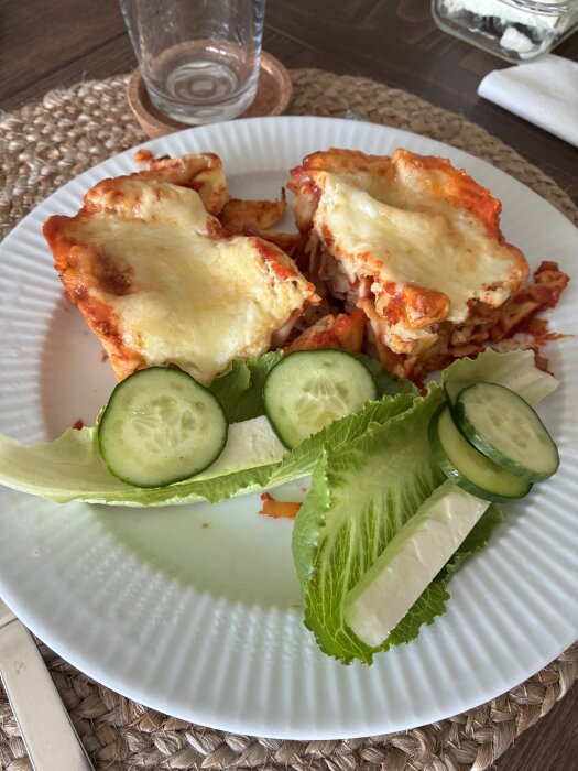 Lasagne med smält ost serverad på en vit tallrik med en sallad bestående av gurkskivor, sallatsblad och oststavar.