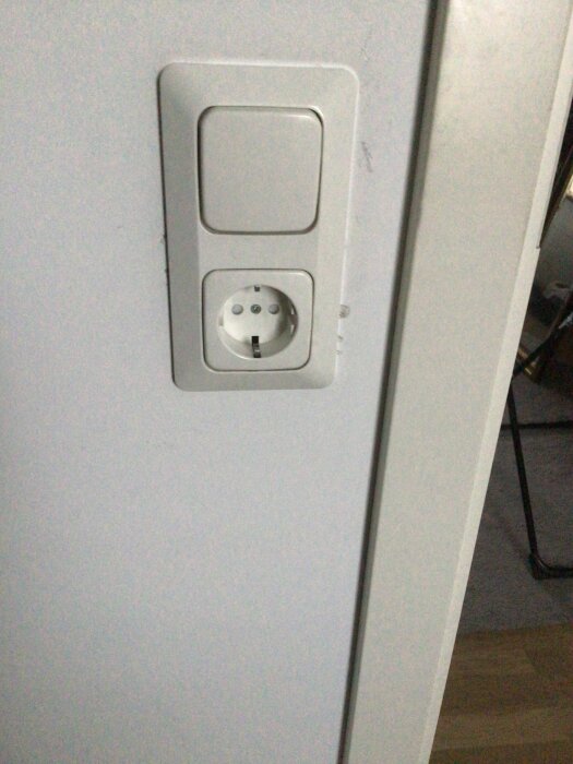 Vägguttag med ett eluttag och en strömbrytare monterade på en vit vägg, använd i diskussion om elinstallation i ett rum.