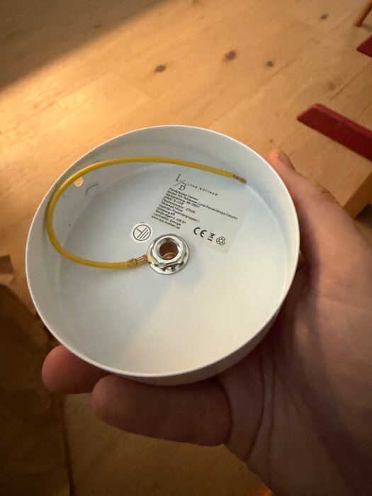 Vit lampbas med gul jordkabel ansluten, hållen i hand. Text och CE-märkning synlig på insidan.
