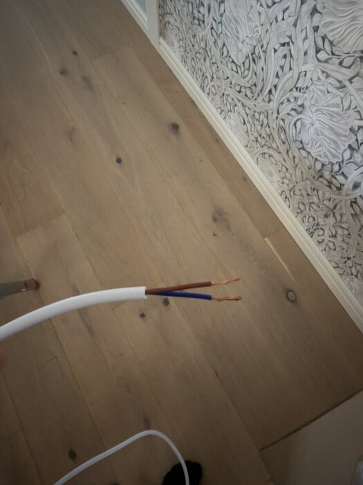 Brun och blå elkabel utan jordledning mot trägolv och tapet.