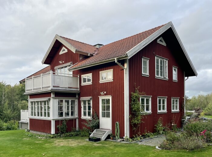 Rött tvåvåningshus med vita fönster och balkong, omgiven av grönska och trädgård.