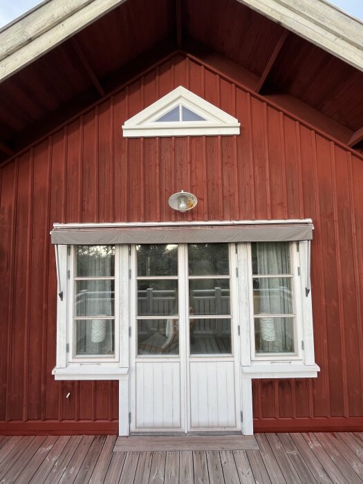 Röd träfasad på ett hus med vita dubbeldörrar och glasfönster, triangelformat fönster högst upp och träterrass framför.