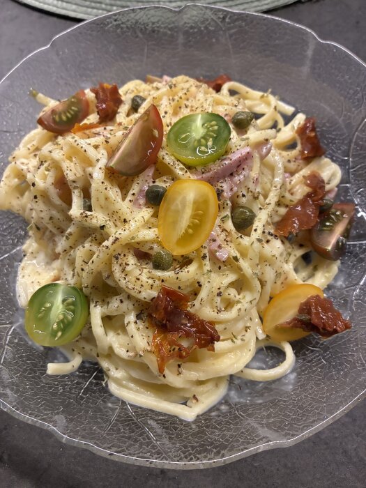 Krämig pasta med cheddar, mozzarella, soltorkade tomater, kapris, skinka och färska tomater toppad med svartpeppar på en genomskinlig tallrik.