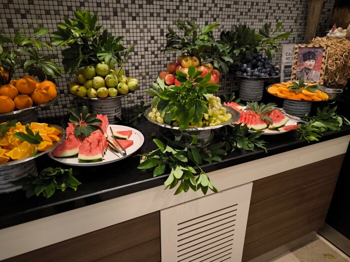 Fruktbuffé med vattenmelon, apelsiner, äpplen, päron, vindruvor och blåbär på tallrikar och fat omgivna av gröna blad.
