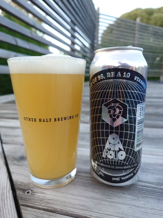 Ett glas ljus öl från Other Half Brewing Co. och en Omnipollo ölburk på ett träbord utomhus.