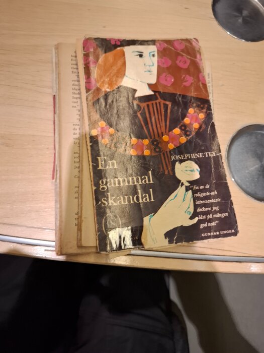 Bok med sliten framsida liggandes på träbord, titeln "En gammal skandal" synlig, skriven av Josephine Tey.