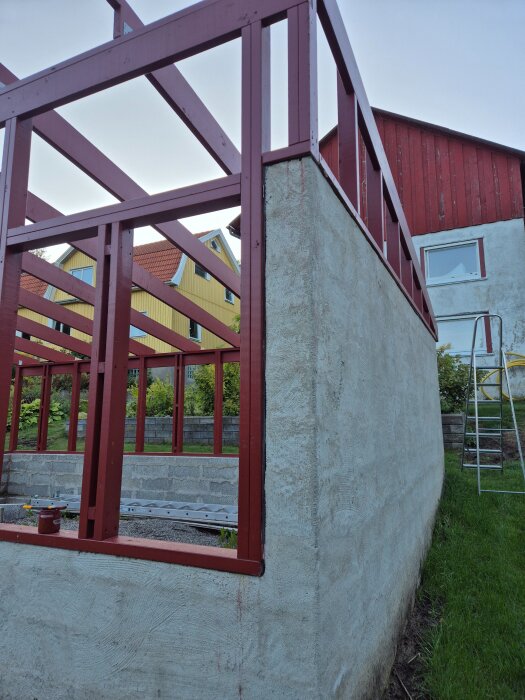 Grundstomme för växthus med röda brädor och rå betongmur; gräsmatta och stege ses intill.