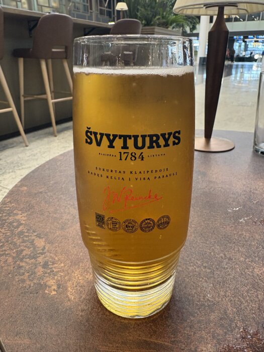 Ett glas Svyturys-öl på ett bord i en flygplatsmiljö, med barstolar och lampor i bakgrunden.