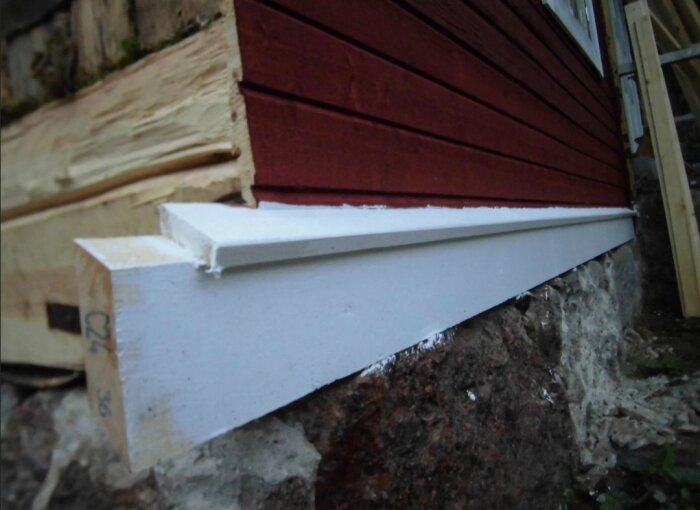 Detaljbild av växthusets nedre del med en vit vattbräda på en röd träfasad och en stenmur som bas.